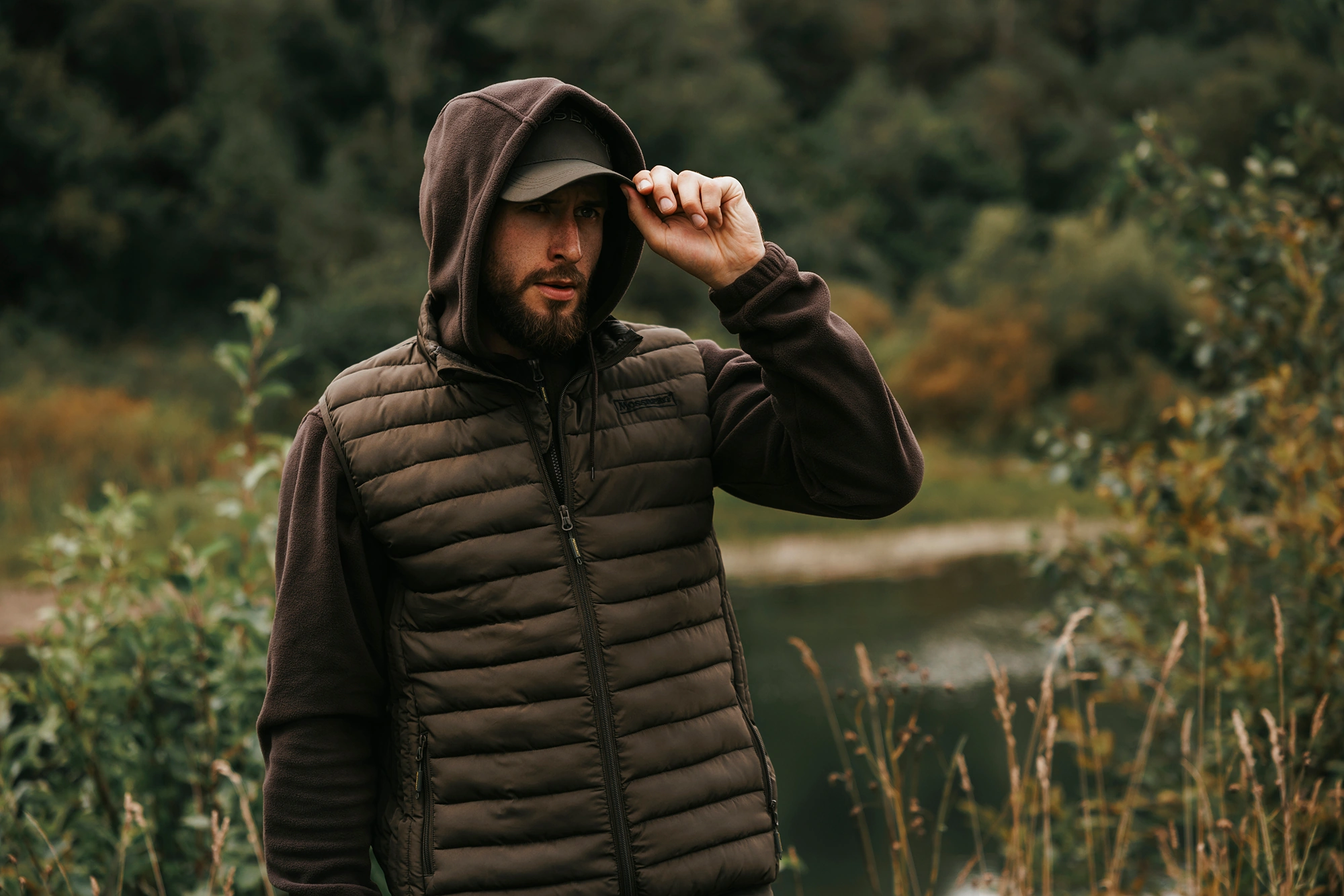 Рыбак в утепленной жилетке поправляет кепку Mossberg.